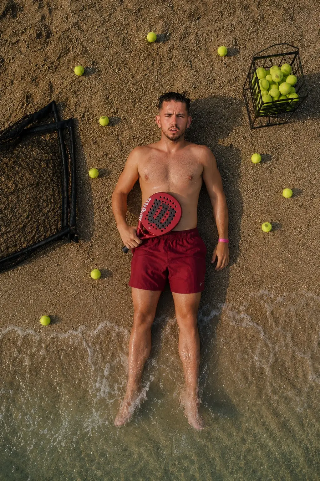 Dani Hoyo tumbado en la orilla del mar con su pala, visto desde arriba, rodeado de pelotas de pádel